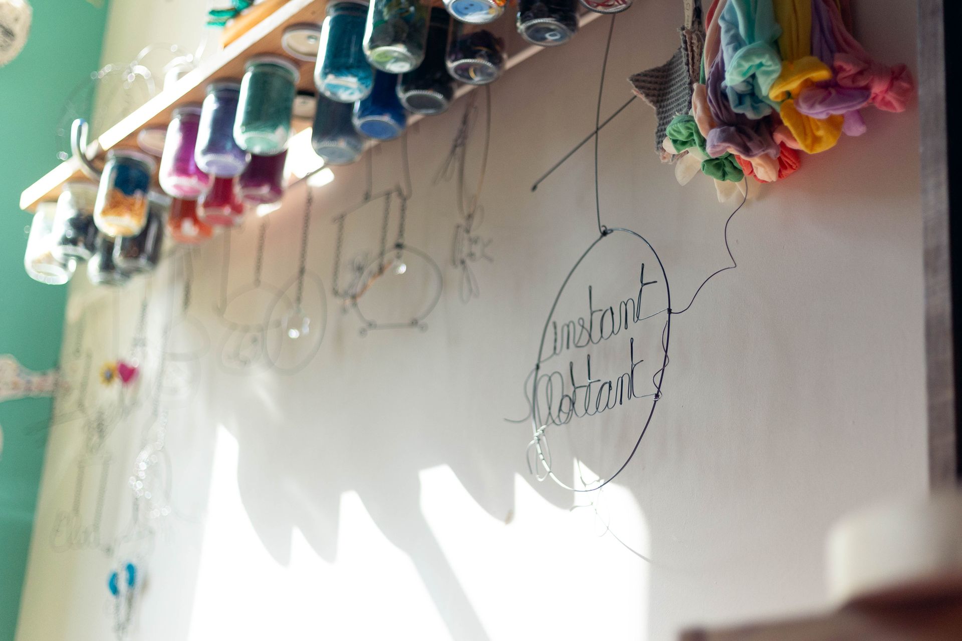 Enseigne en fil de fer « Instant Flottant » suspendue dans l'atelier, pots de laine multicolores accrochés à l'envers, tissus colorés et mobiles en fil, jeu d'ombre et de lumière sur le mur